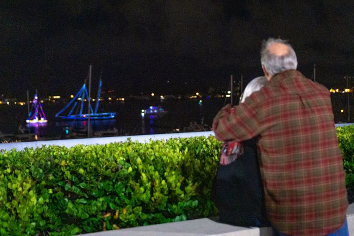 A couple looks onto the bay and a few boats are floating on the water and lit up with lights.